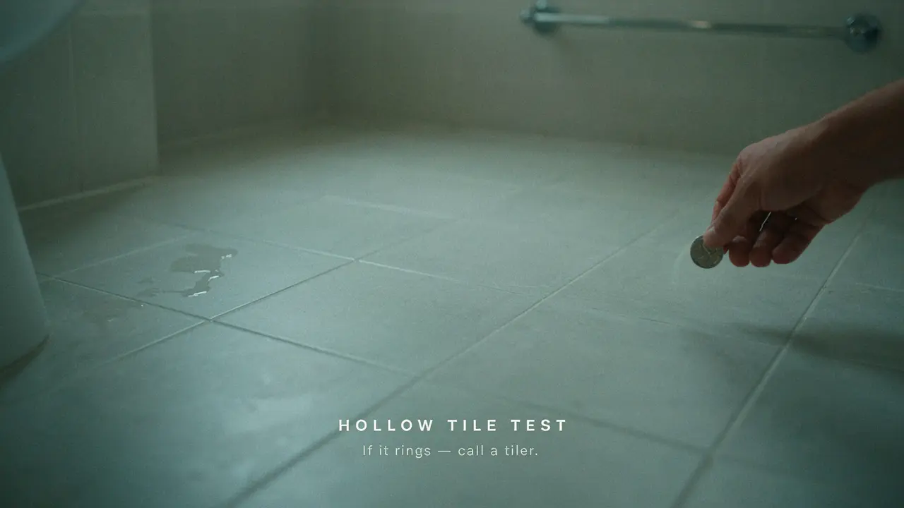 Person tapping a bathroom floor tile with a coin to test for hollow sound indicating failed adhesive bond in a Melbourne home